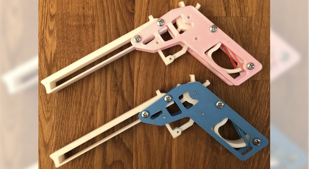 two 3d printed rubber band guns, one in pink and one in blue, displayed on a wooden surface. The guns have a rotating drum mechanism that allows for semi-automatic 6 shot action.