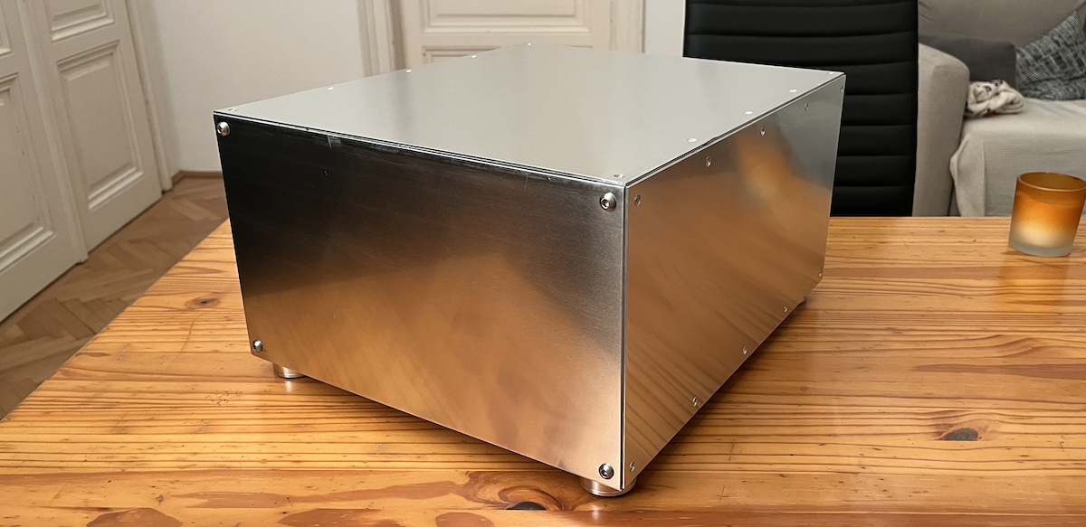 A custom silent home server case built from brushed aluminium sheets, sitting on a wooden table. The case has a clean rectangular shape with visible screws at the corners and small aluminium HiFi-style feet.